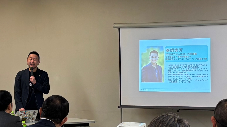 講師が講演をする様子を部屋の後方から写した写真。マイクを持った講師と、ホワイトボードに自己紹介スライドが映っている。